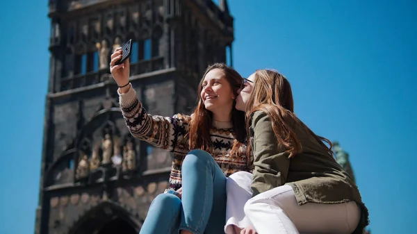 Dos mujeres jóvenes se sientan debajo de la vieja torre y se toman una selfie con una sonrisa —  Fotos de Stock