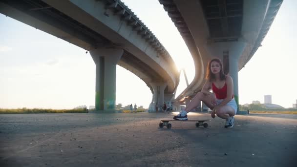 Young attractive woman sitting near the skateboard under the bridge - bright sunlight — Stock Video