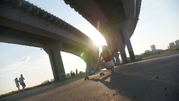 Fiatal vonzó nő ül a gördeszka alatt a híd-ragyogó napfény — Stock videók