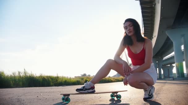 Young attractive woman sitting near the skateboard under the bridge - bright sunlight on a sunset — Stock Video