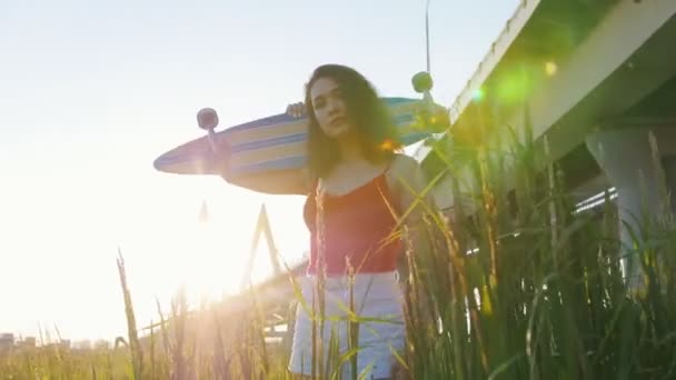 Jovem mulher atraente com skate em pé na grama alta e posando para a câmera — Vídeo de Stock