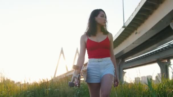 Jovem mulher em roupas de verão andando entre grama alta segurando um skate - pôr do sol - ponte urbana — Vídeo de Stock