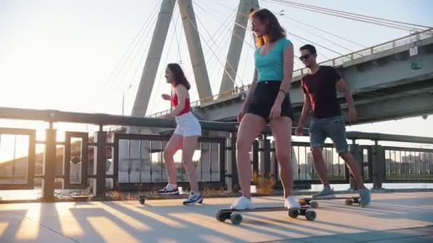 Молодые улыбающиеся друзья катаются на скейтбордах по набережной - закат — стоковое видео