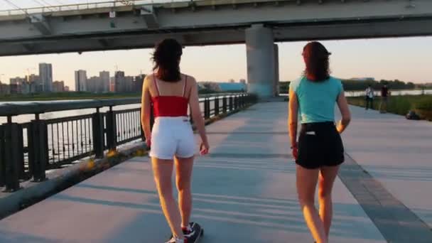 Молодые женщины катаются на скейтбордах по набережной под мостом — стоковое видео
