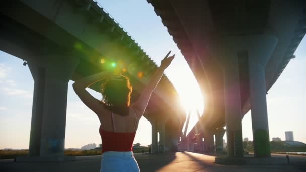 Mujer joven en pantalones cortos blancos y camisa roja montando monopatín bajo el puente urbano y agitando las manos - luz del sol brillante — Vídeo de stock