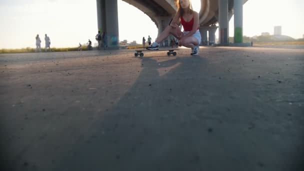 Mujer atractiva joven sentada en el monopatín bajo el puente urbano - luz solar brillante — Vídeos de Stock