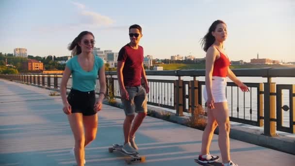 Юные друзья катаются на скейтбордах по набережной на фоне современных зданий - желтый закат — стоковое видео