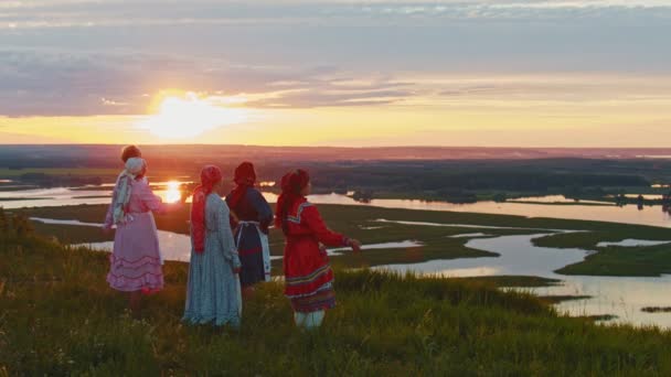 Νέοι με ρωσικά παραδοσιακά ρούχα που στέκονται στο χωράφι και απολαμβάνουν τη θέα στο ηλιοβασίλεμα-ποταμό και τα μικρά νησιά — Αρχείο Βίντεο