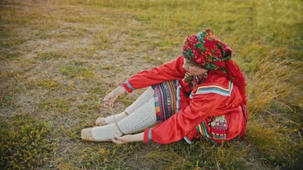 Eine Frau in russischer roter Volkskleidung zieht Bastschuhe an - sitzt auf dem Feld — Stockvideo