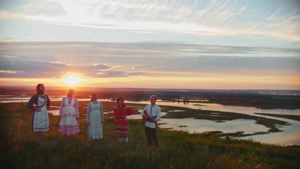 Jonge mensen in Russische traditionele kleren staan op het veld op een achtergrond van prachtige zonsondergang en zingen — Stockvideo