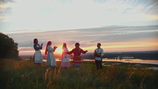 Młodzi ludzie w rosyjskich tradycyjnych ubrań zabawy na boisku na tle pięknego zachodu słońca-człowiek grający Balalaika i kobiety tańczą przez muzykę — Wideo stockowe
