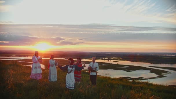 Jovens em roupas tradicionais russas se divertindo no campo em um fundo de belo pôr do sol - cantando, dançando — Vídeo de Stock