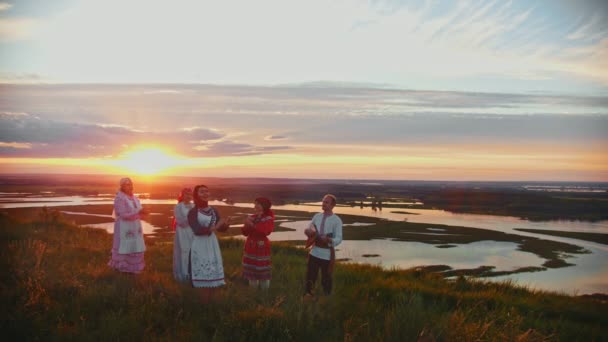 Jonge mensen in Russische traditionele kleren staan op het veld op een achtergrond van prachtige zonsondergang en zingen — Stockvideo