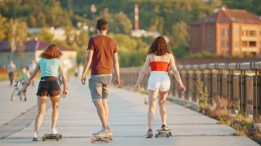 Gün batımında modern binaların arka planında iskelede kaykay lar süren iki kadın ve bir adam