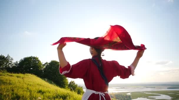 Mladá žena v ruském tradičním oblečení, která kráčí po zeleném poli-její červený kapesník se chvěje větrem