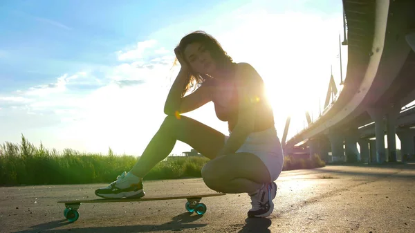 Jeune femme séduisante assise sur le skateboard sous le pont - lumière du soleil éclatante — Photo