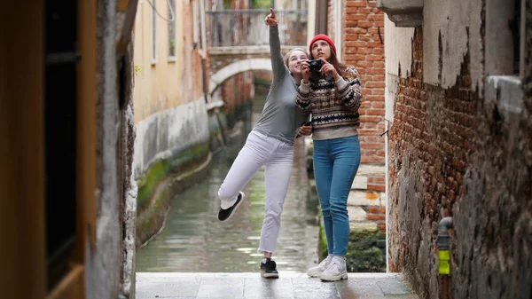 Duas mulheres jovens viajantes em roupas quentes em pé sobre um fundo de um canal de água e tirar fotos - Veneza, Itália — Fotografia de Stock