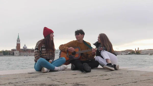 Skupina mladých přátel, kteří sedí na kraji přístaviště a zpívají písně u kytary-Benátky, Itálie — Stock fotografie