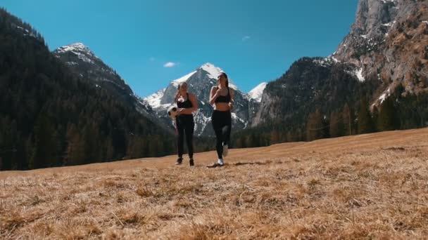 Duas jovens mulheres em sutiã esportivo correndo em direção à câmera segurando uma bola de futebol - Dolomites, Itália — Vídeo de Stock