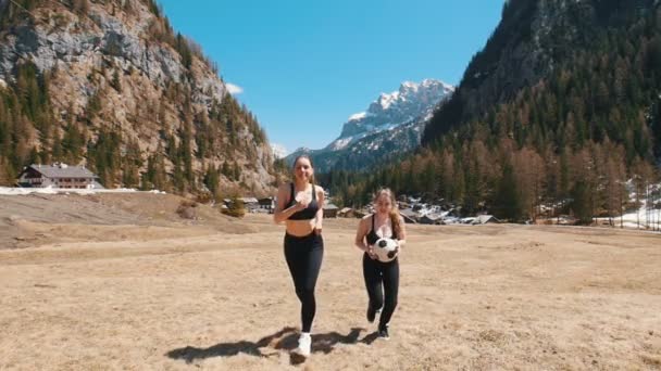Две молодые женщины бегут к камере, держа футбольный мяч на фоне леса - Доломиты, Италия — стоковое видео