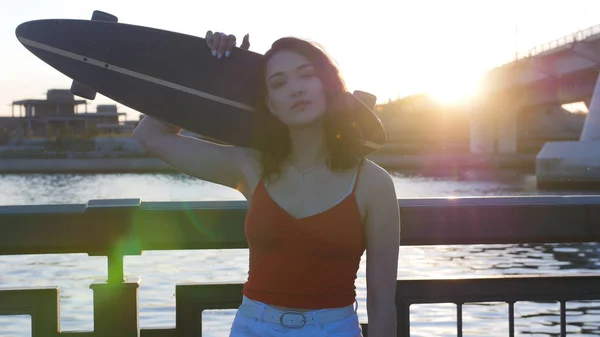 Νέα γυναίκα στέκεται στην προκυμαία στο ηλιοβασίλεμα κρατώντας ένα σκέιτμπορντ-ποζάρει στην κάμερα — Φωτογραφία Αρχείου