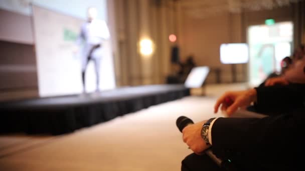 En man i den blå kostym talar på scenen-Business konferens i hallen-en annan man sitter på stolen och läsa dokument som håller en mikrofon — Stockvideo