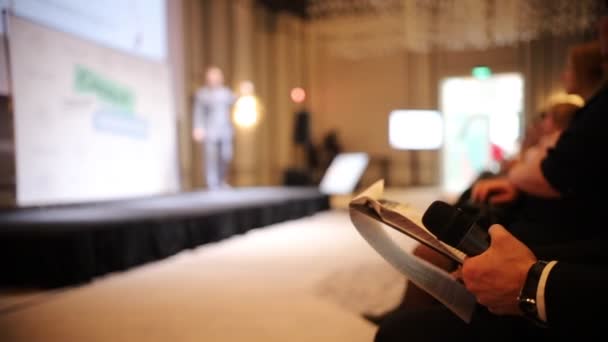 Чоловік у блакитному костюмі говорить на сцені - бізнес-конференція в залі - інший чоловік сидить на стільці і читає документи — стокове відео