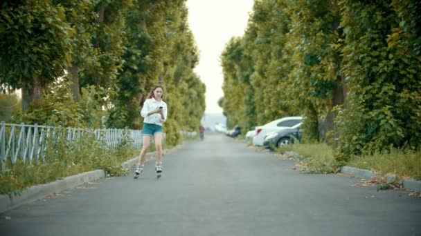 一个十几岁的女孩在轮滑轮上溜冰的道路上 - 看着电话 — 图库视频影像
