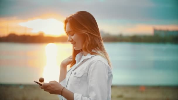 En leende tonårs flicka stående på vattnet-titta på telefonen-hålla en klubba-solnedgång — Stockvideo