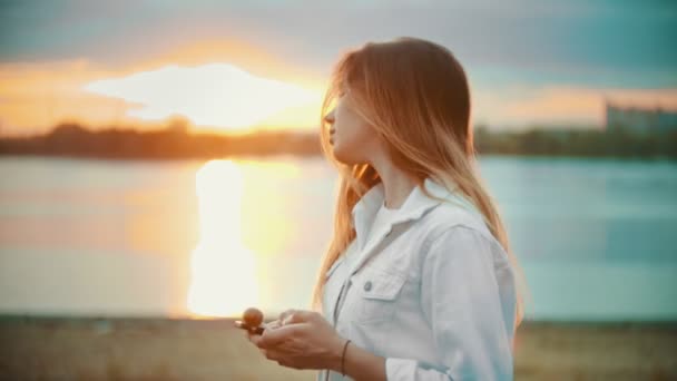 Дівчинка-підліток стоїть на набережній дивиться на телефон тримаючи льодяник захід сонця — стокове відео
