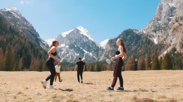 Молодые друзья играют в футбол и бадминтон на поле на фоне гор и лесов — стоковое видео