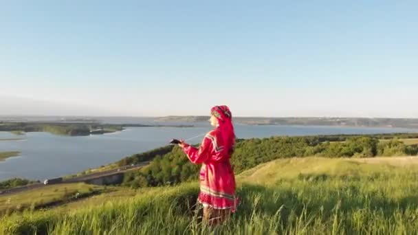 緑の丘に立ってバラライカを再生する伝統的なロシアのドレスを着た若い女性 — ストック動画