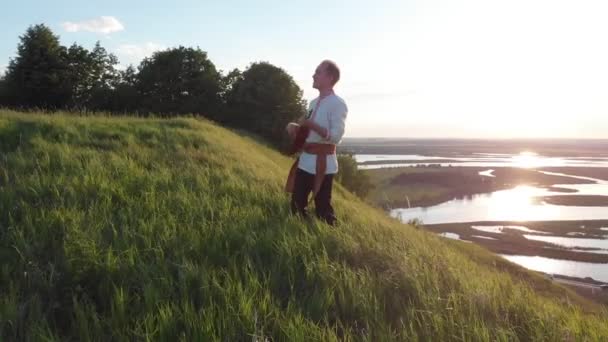 緑の丘に立って、バラライカを再生する伝統的なロシアの服を着た若い男 - カメラは、その周りを撮影. — ストック動画