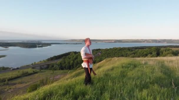 Jeune homme en vêtements traditionnels russes debout dans l'herbe à la colline verte et jouer balalaika - Coucher de soleil — Video