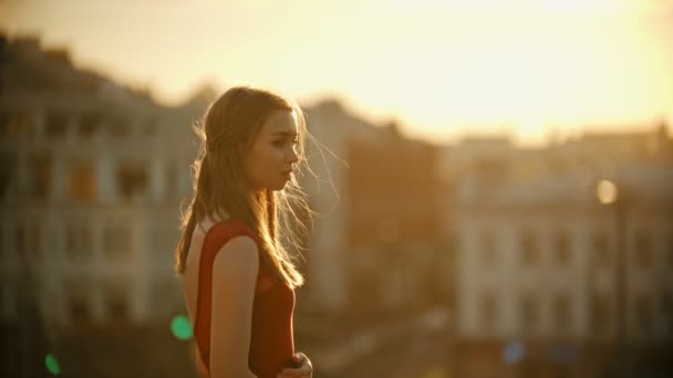 Молодая женщина в красном платье стоит на крыше на закате — стоковое видео