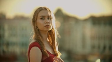 Kırmızı elbiseli genç bir kadın çatıda duruyor ve gün batımında kameraya poz vermek.