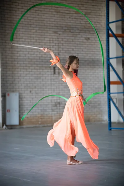 Joven mujer en forma de vestido naranja bailando con cinta gimnástica en las manos en el interior —  Fotos de Stock