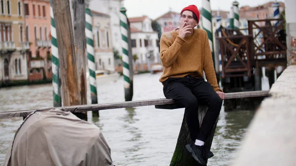 Een jonge man zit op de Bank boven het water en rookt — Stockfoto