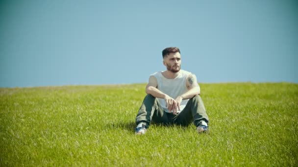 Egy fiatal férfi tetoválással ül egy ragyogó zöld fű — Stock videók
