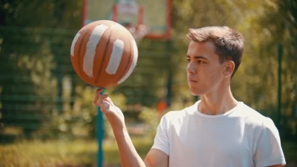 Молодий красивий чоловік стоїть на спортивному майданчику і обертається баскетбольним м'ячем на пальці — стокове відео
