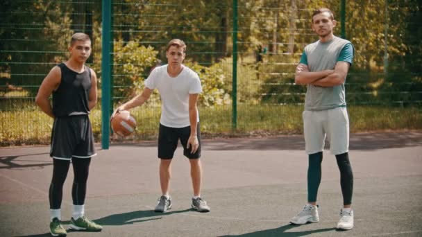 三个运动员站在室外的篮球场上，看着镜头——一个运动员把球从地上打下来 — 图库视频影像