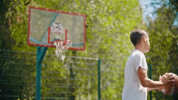 Молодой человек стоит на спортивной площадке и бросает мяч в баскетбольное кольцо - попадает в цель — стоковое видео