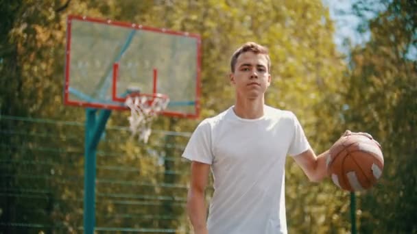 Un joven en camiseta blanca de pie en un campo de deportes y descarta la pelota del suelo — Vídeos de Stock