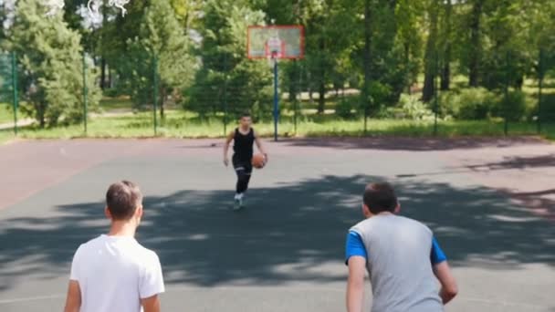 屋外のコートでバスケットボールをしている3人のスポーツマン - 黒いユニフォームを着た男がボールを投げる — ストック動画