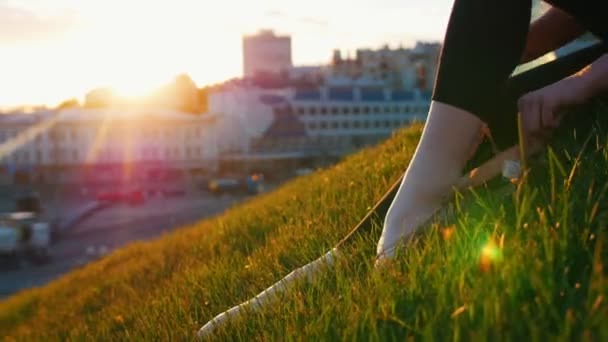 Junge attraktive Ballerina zieht ihre Spitzenschuhe aus, sitzt auf einem grünen Hügel mitten in der Stadt. — Stockvideo