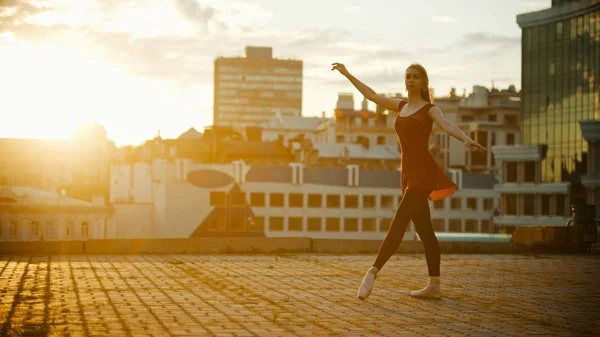 Молода витончена жінка в червоній сукні балерини, що стоїть в позі на даху яскравий захід сонця — стокове фото