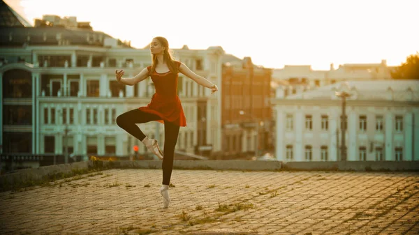Fiatal ihletett nő piros ruhában balerina állt a kecses jelent a tető-modern épület a háttérben-fényes naplemente — Stock Fotó