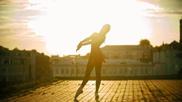 Une silhouette de jeune femme ballerine en robe debout dans la pose gracieuse sur le toit coucher de soleil lumineux — Photo