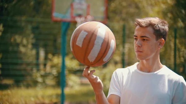 白いシャツを着た若いハンサムな男がスポーツ場に立ち、バスケットボールのボールを指で回転させる — ストック写真