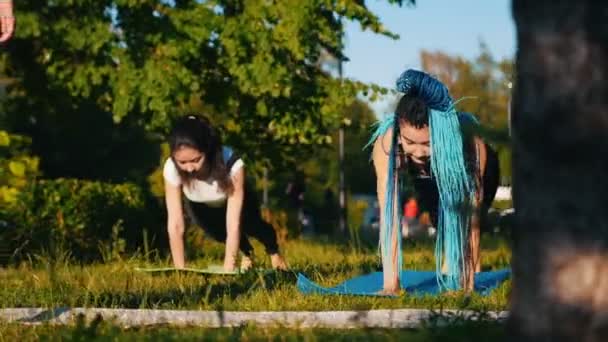 Två unga kvinnor som gör yoga övningar med instruktör i parken-en kvinna har långa blå dreadlocks — Stockvideo